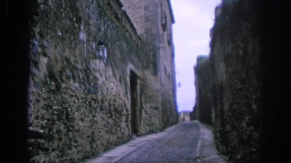 1966 Spain 8mm frame showing a narrow cobbled street bordered by weathered stone walls and a mounted lantern.