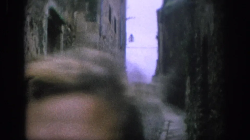 Soft 8mm frame with a cropped young face near the lens and a cobbled stone lane receding in the background.