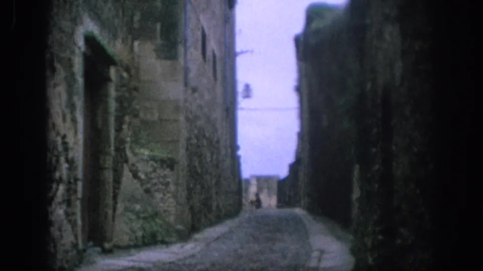 1966 Spain home-movie frame of a narrow cobbled street bordered by tall stone walls and distant figures.