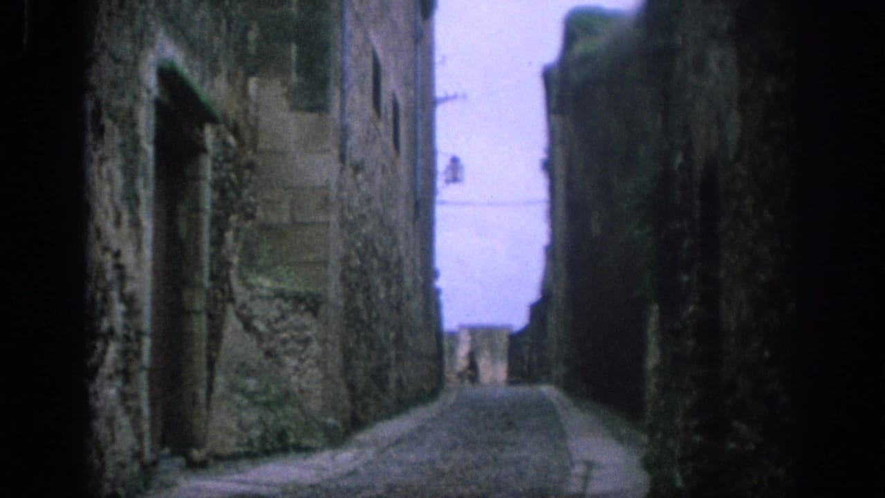 1966 Spain 8mm frame of a narrow paved alley between rough stone walls, with an old window and a distant person near the far end.