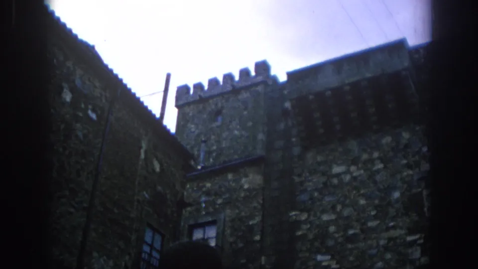 1966 Spain 8mm frame looking up at fortress-like stone walls, crenellated battlements, a small window, and overhead utility wires.