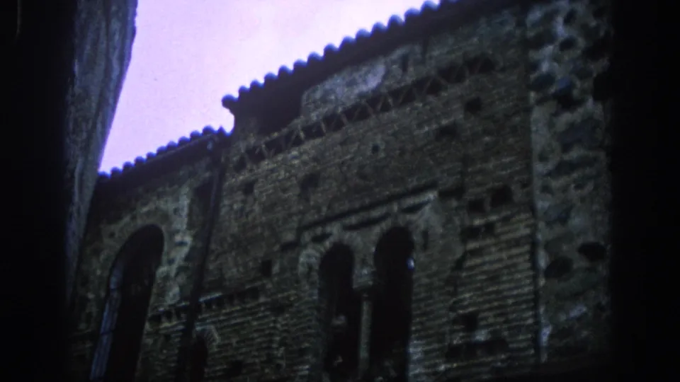 1966 Spain home-movie frame of a weathered brick building with arched window openings and roof tiles, shot upward from a narrow street.