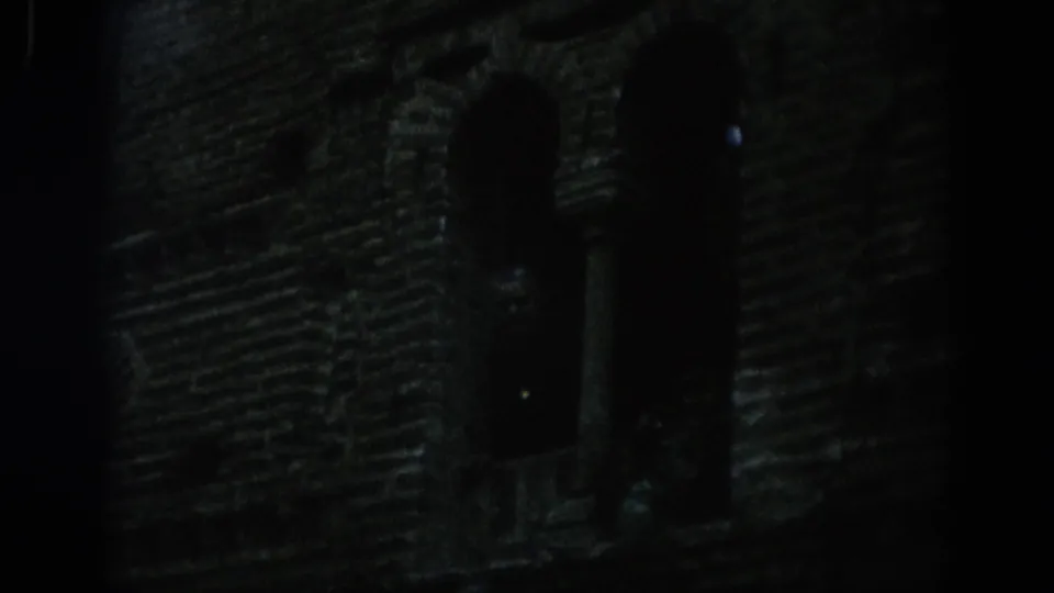 A dim 1966 Spain 8mm frame isolates an arched window opening in a weathered historic masonry facade.