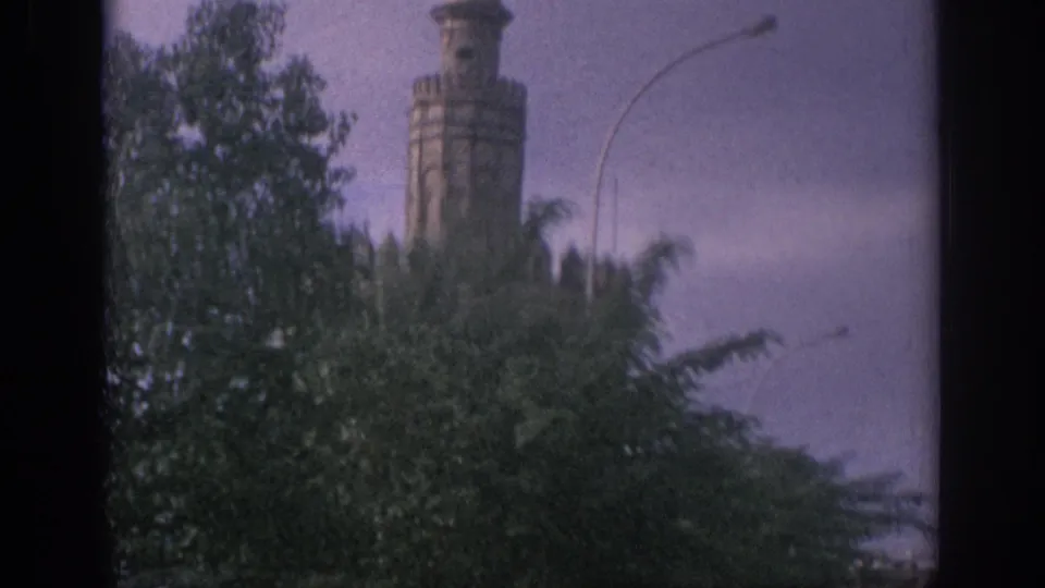 1966 Spain home-movie frame showing a stone tower rising behind dense trees on an urban street.