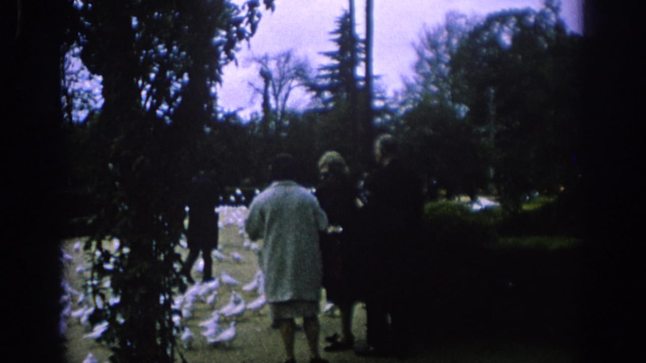 In 1966 Spain, an older woman in a light coat appears to feed white doves while bystanders stand in a shaded park.