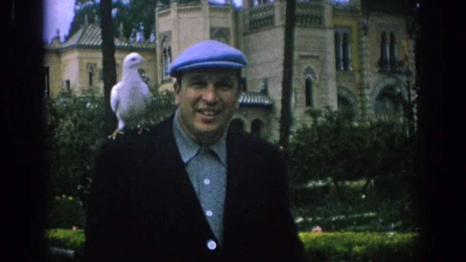 In the 1966 Spain home movie, a man in a blue cap faces the camera with a white dove on his shoulder before an ornate park building.