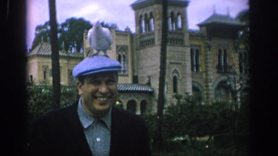 In Spain 1966, a white dove perches on a smiling man’s blue cap with garden foliage and an ornate building behind him.
