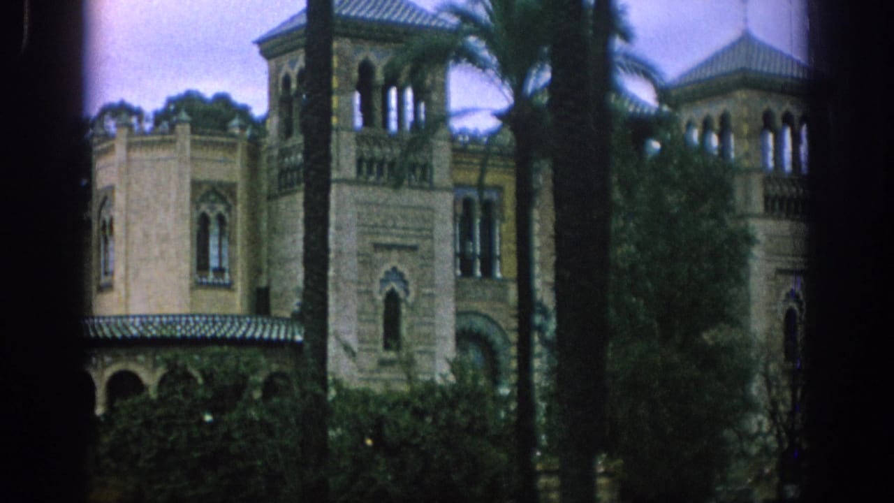 1966 Spain home-movie view of an ornate tan building with towers, arched windows, palms, and dark foreground vegetation.