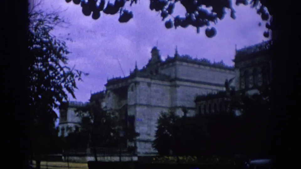 1966 Spain home-movie frame showing a darker view of a pale building partly hidden by trees, with silhouetted foliage and blue-purple color shift.