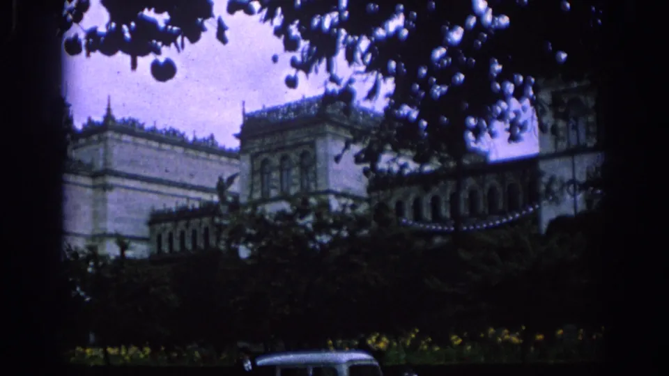 A Spanish architectural exterior appears behind trees and garden foliage, with a brief mid-century car roof visible near the lower edge.