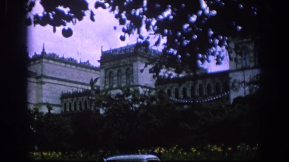 Purple-tinted 8mm frame of a Spanish building with tower details, seen from a shaded garden area through dense foreground foliage.