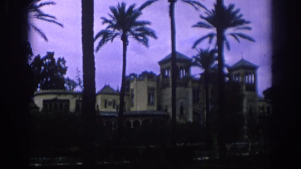 Palm trees frame a Spanish historic-looking exterior with towers and arched details, captured in soft purple-tinted 8mm color.
