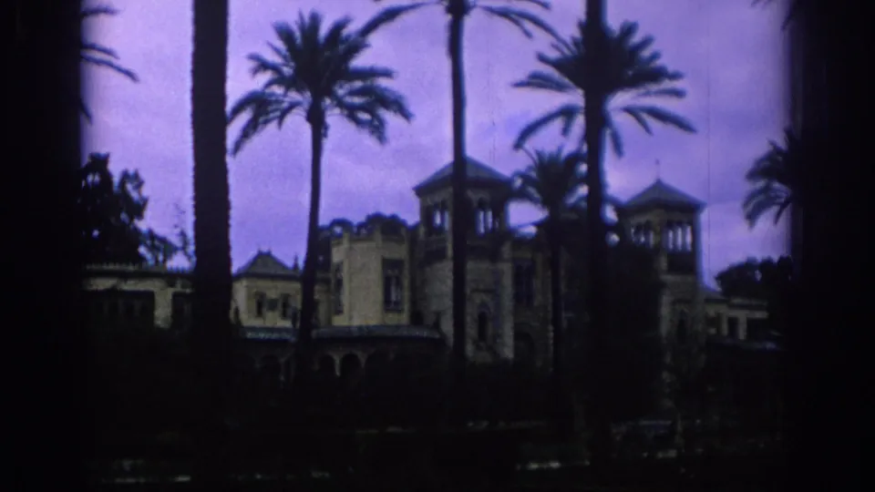 1966 Spain home-movie frame showing a palm-lined resort-like building in soft, dark 8mm color.