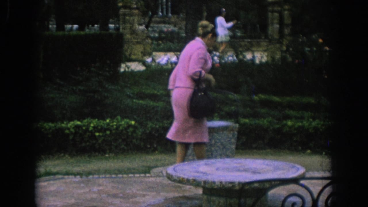 In Spain, 1966, a woman in a pink dress carries a black handbag through a formal park garden.
