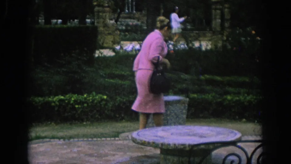 In Spain, 1966, a woman in a pink dress carries a black handbag through a formal park garden.
