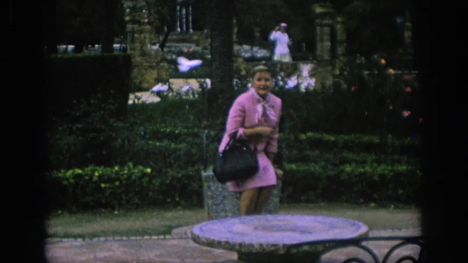 In Spain, 1966, a woman in a pink dress pauses by a stone bench as pigeons move in the background.