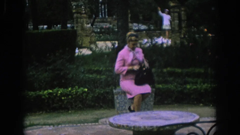 In Spain, 1966, a woman in a pink dress sits or poses on a stone bench with pigeons and hedges behind her.