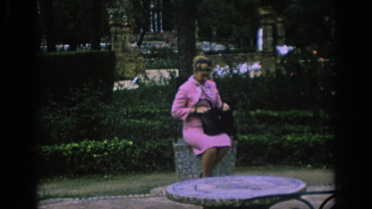 Woman in a pink suit sits on a stone bench in a leafy garden square.