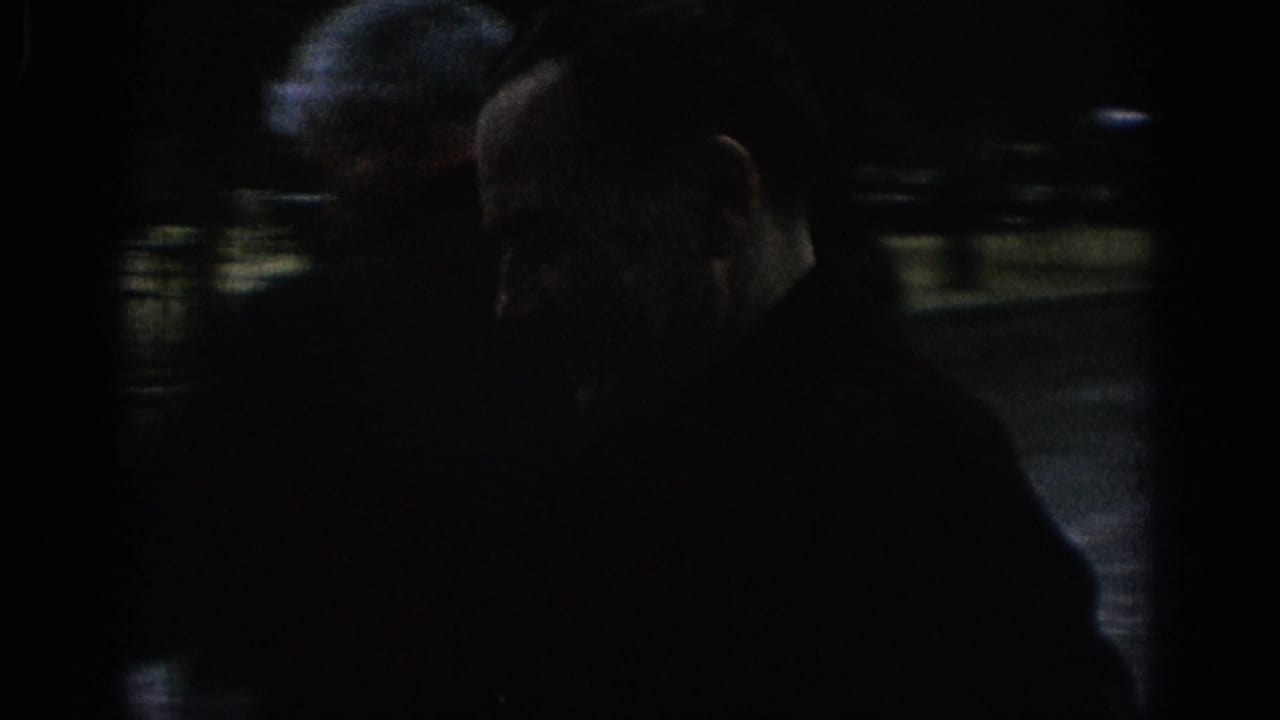 Very dark close view of an adult man near a street, with another figure partly visible behind him.