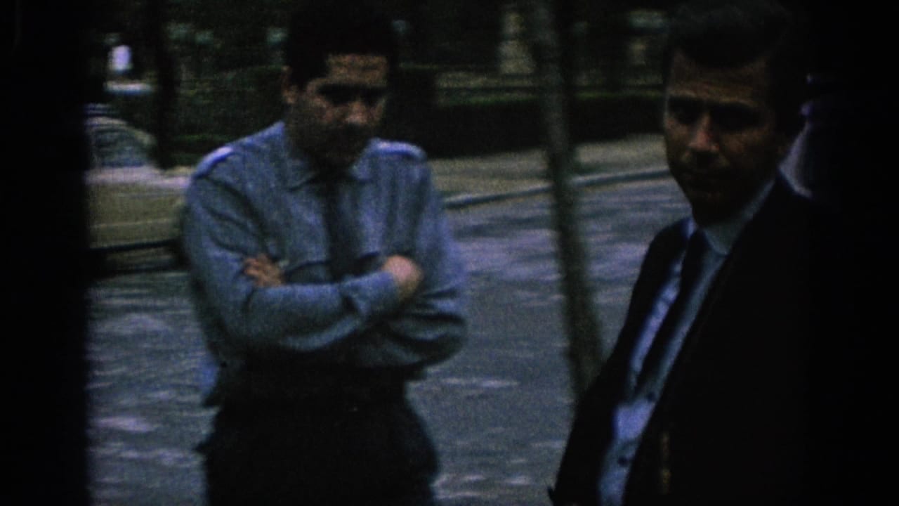 Two adult men stand on a city street, one in a light shirt with arms crossed and one in a dark suit.