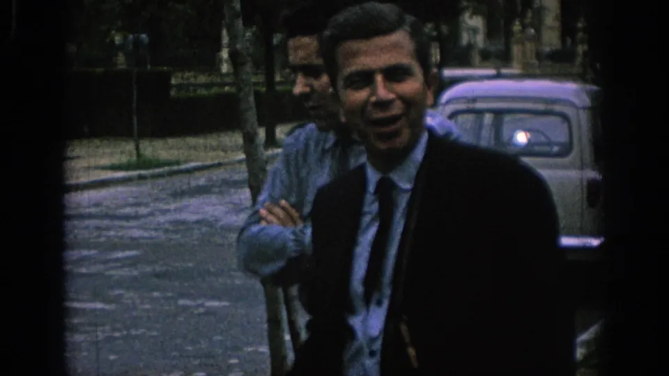 1966 Spain home-movie frame showing a smiling suited man in front of another man, with a rounded car parked behind them.