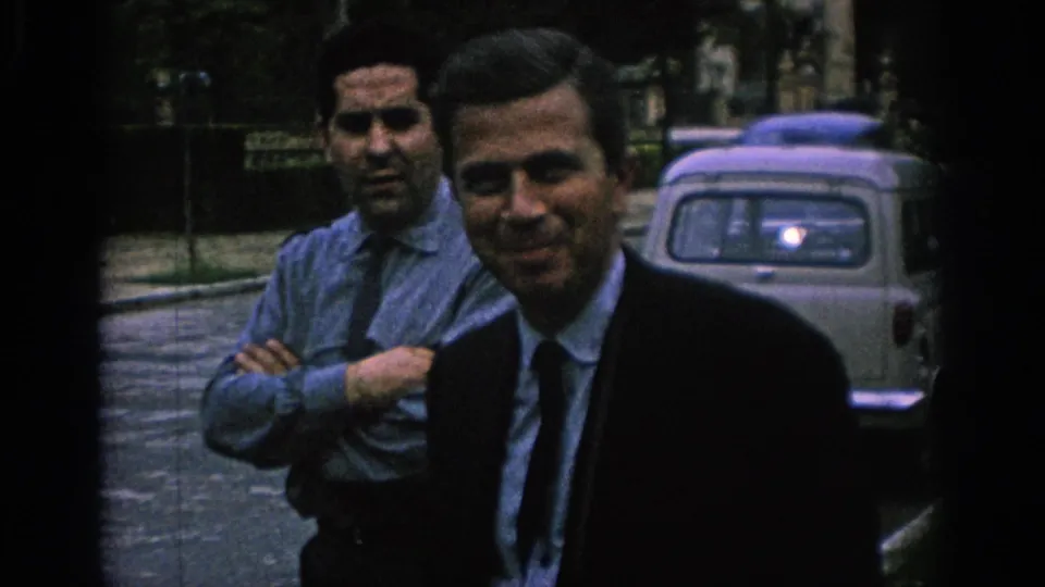 1966 Spain street frame with two adult men facing the camera, one in a dark suit and tie and one in a light shirt, with a car behind.