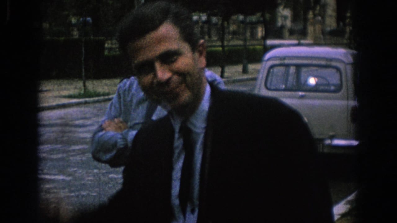 In 1966 Spain footage, a suited adult man smiles and waves near a pale vintage car as another adult man stands with arms crossed.