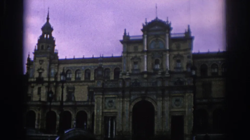 1966 Spain home-movie frame of Plaza de Espana showing the large central arch, decorative facade, tower, and paved courtyard foreground.