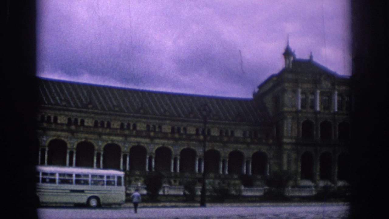 Long colonnaded building with a white bus parked in front and one person crossing the plaza.