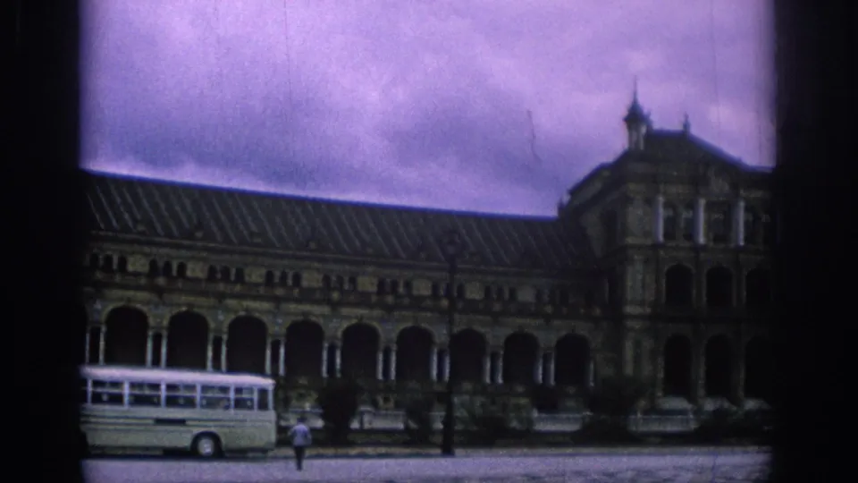 1966 Spain travel footage frame with a white tour bus, lone pedestrian, arched colonnade, and ornate building wing.