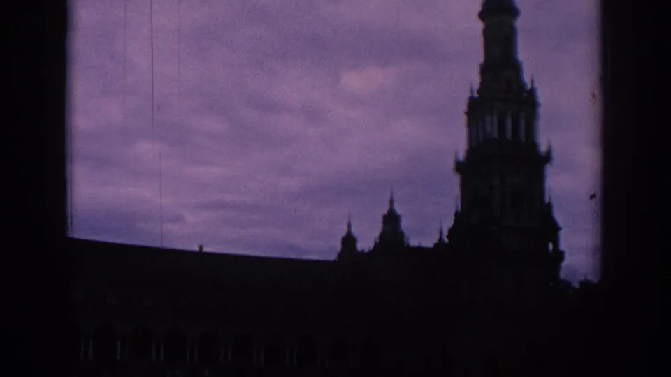 1966 Spain 8mm frame with a shadowed ornate building, tall spire, smaller rooftop details, and violet clouds behind it.
