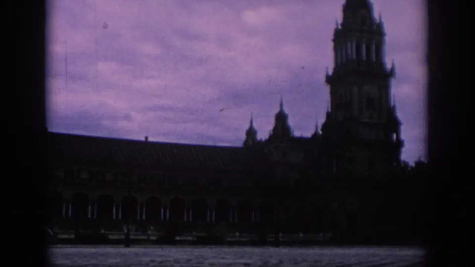 1966 Spain 8mm frame revealing more of the towered structure, including a dark colonnade and open paved area below the violet sky.