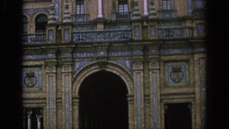 1966 Spain home-movie frame centered on a large arched entrance beneath carved stonework, balcony rails, tile panels, and dark gate openings.