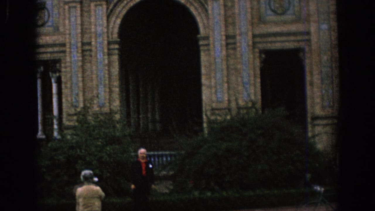 1966 Spain home-movie frame showing an ornate arched facade, dark entryways, shrubs, and distant visitors in a courtyard.
