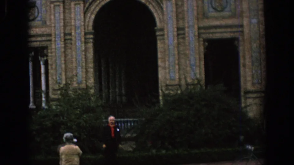 1966 Spain home-movie frame showing an ornate arched facade, dark entryways, shrubs, and distant visitors in a courtyard.
