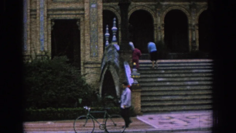 1966 Spain home-movie frame showing visitors on broad stairs, a bicycle below, shrubs, railings, and arched architectural details.