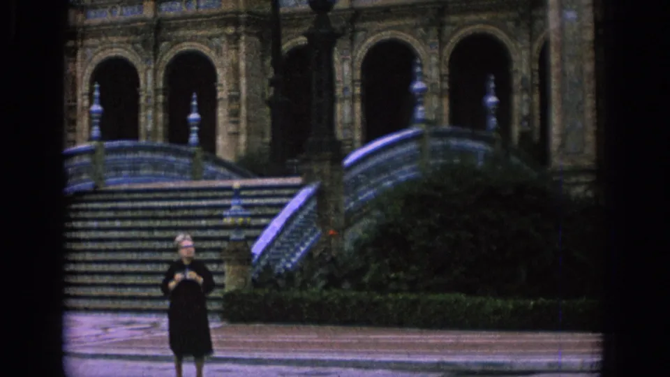 1966 Spain frame with a person standing in the courtyard before wide stairs, curved railings, shrubs, and ornate arched entrances.