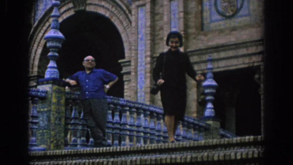 1966 Spain home-movie frame of the woman moving along the balcony as the man poses near the arched entrance.