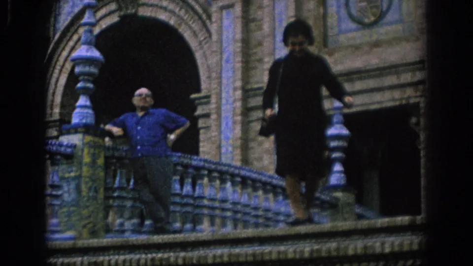 In 1966 Spain home-movie footage, a man stands at a blue and yellow tiled balustrade while a woman in dark clothing descends the stairs.