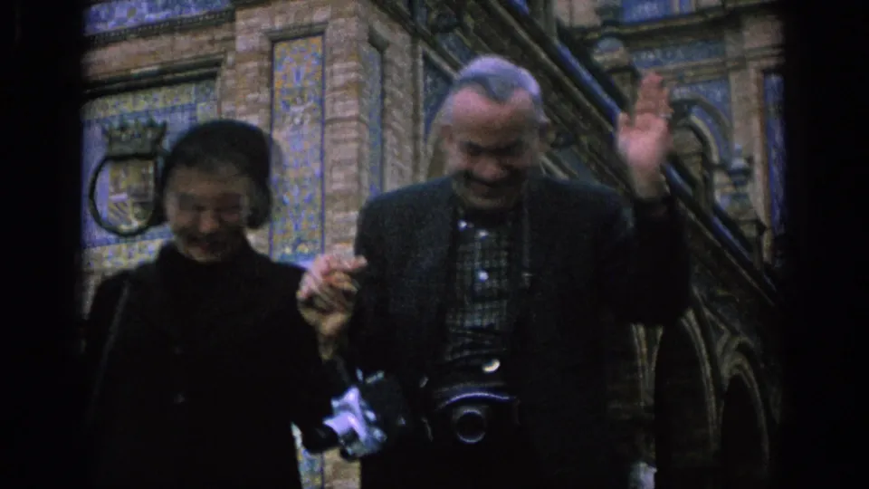 In Spain in 1966, a smiling couple walks near ornate tilework and stairs while the man waves toward the 8mm camera.
