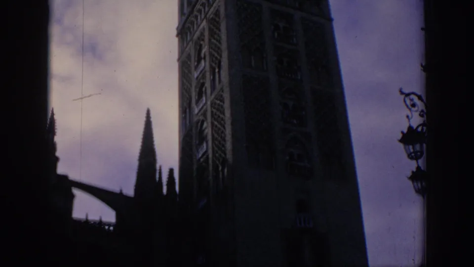 1966 Spain 8mm frame showing a tall historic tower in daylight, with shadowed foreground architecture and a decorative street lamp.