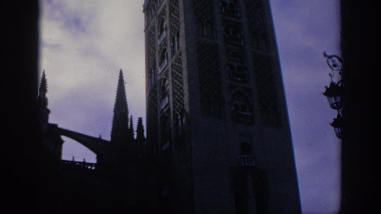 1966 Spain 8mm frame showing a shadowed low-angle view of a Gothic cathedral tower, pointed spire, and cloudy sky.