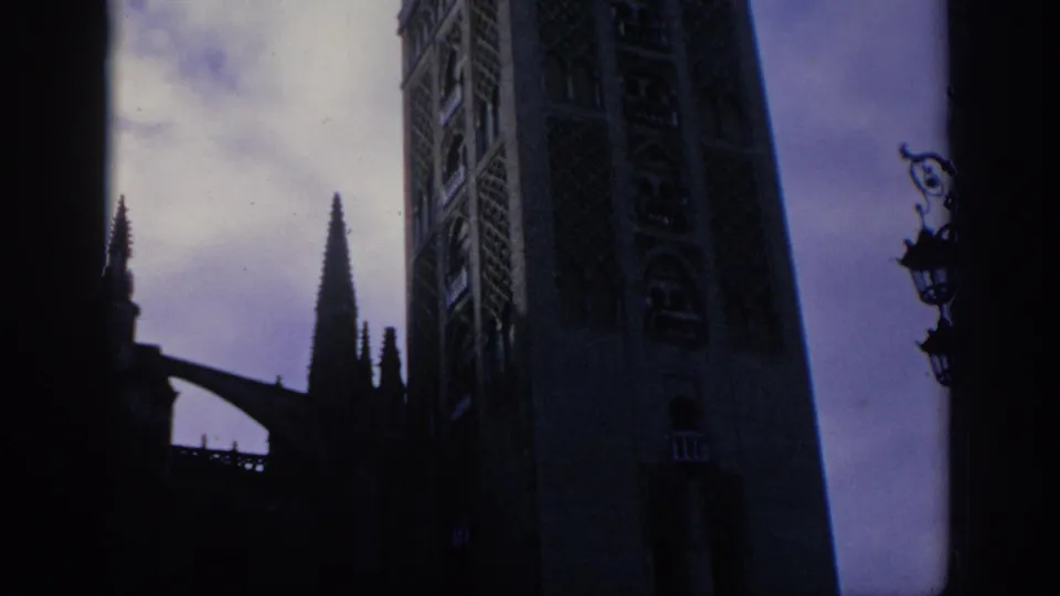 1966 Spain 8mm frame showing a shadowed low-angle view of a Gothic cathedral tower, pointed spire, and cloudy sky.
