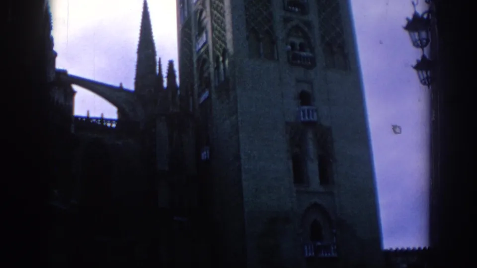 1966 Spain 8mm frame showing a Gothic cathedral tower rising past dark foreground shapes under a cloudy sky.