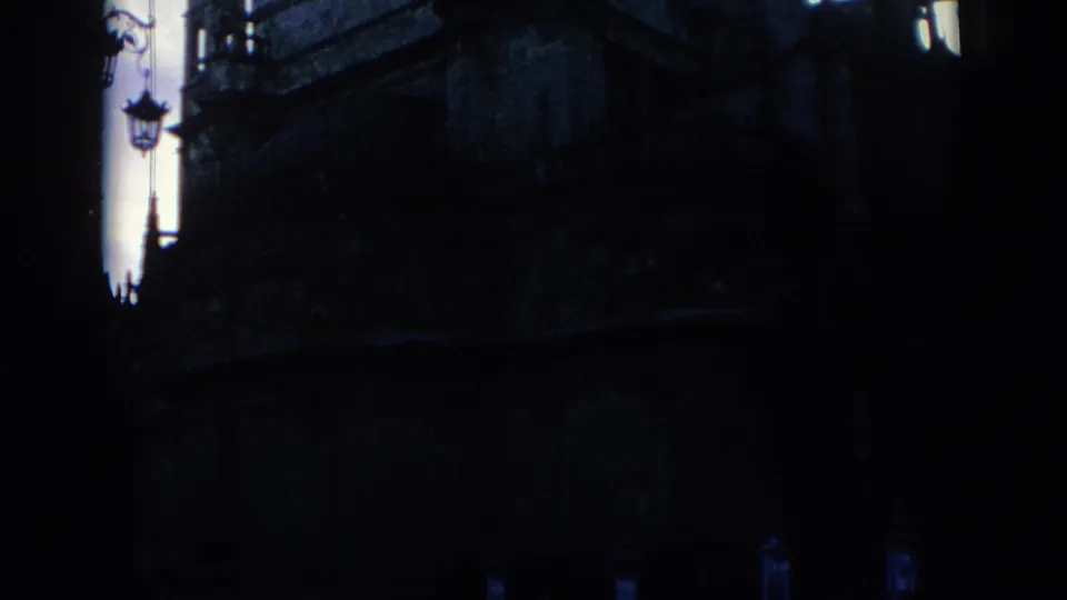 1966 Spain home-movie frame looking from deep shade toward a historic stone exterior, with a decorative streetlamp visible at left.