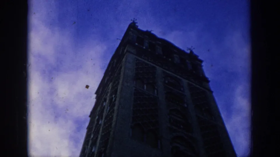 A dark cathedral-like Gothic tower in Spain rises against a blue-purple dusk sky in grainy 1966 8mm footage.