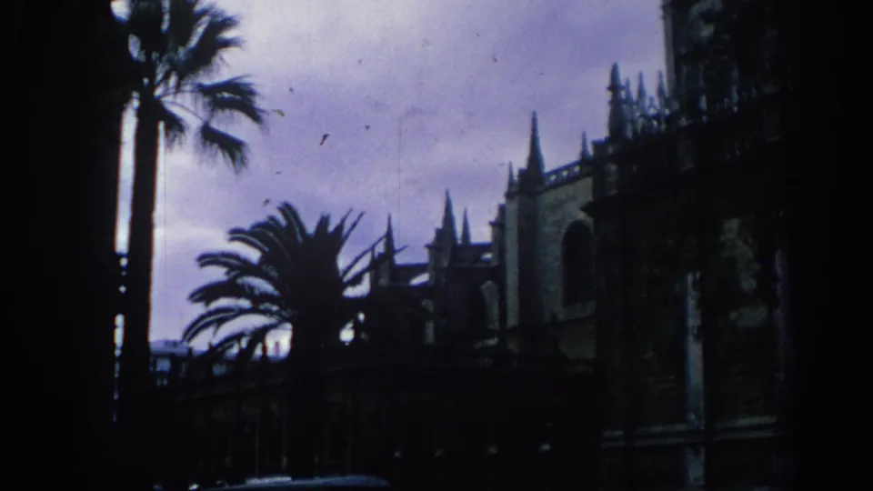 1966 Spain 8mm frame showing palm trees and the shadowed exterior of Seville Cathedral under a dim sky.