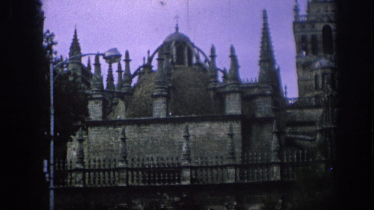 1966 Spain 8mm frame showing an ornate historic exterior with spires, domes, a tower, fence line and purple-tinted sky.