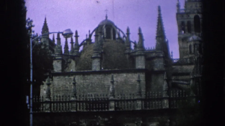 1966 Spain 8mm frame showing an ornate historic exterior with spires, domes, a tower, fence line and purple-tinted sky.