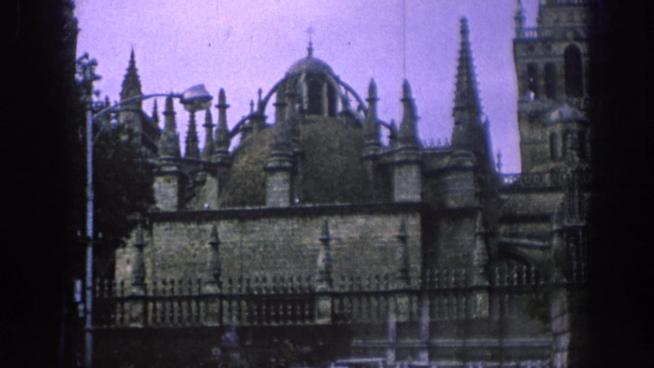 Similar view of a spired stone facade with domes, tower, fence and streetlamp.